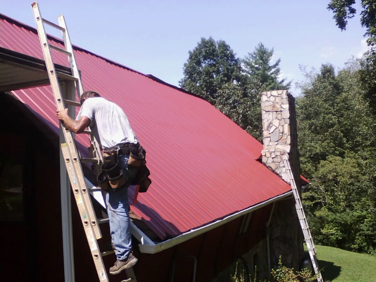 Roofer on a ladder beside a red metal roof and stone chimney — Soffit & Fascia Repair by Roofing Brothers in Buckner